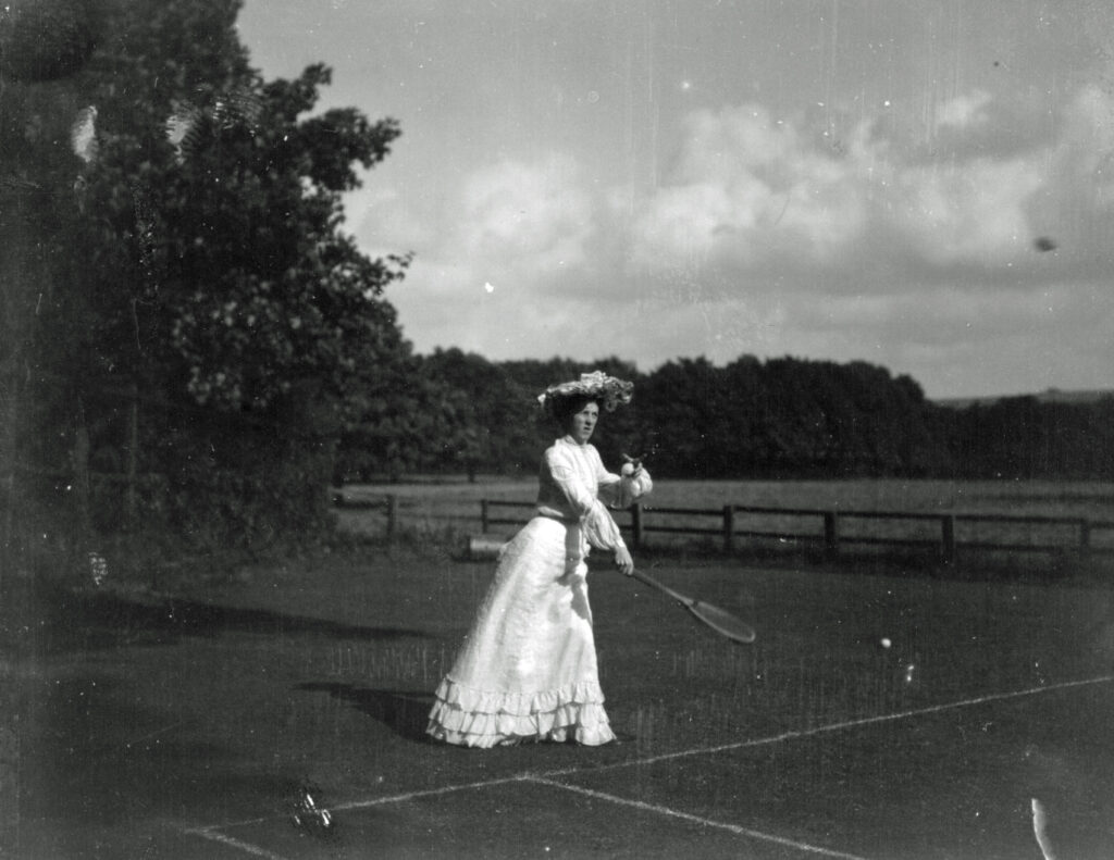 Lady tennis player with hat and long skirt serves in tennis match